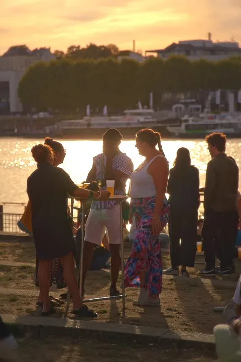 Des spectateurs discutent autour d’un mange-debout, un verre à la main, face à la Loire éclairée par le soleil couchant.