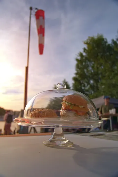 Une cloche en verre protège un burger végétal devant une manche à air rouge et blanche, au coucher du soleil.
