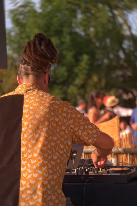 Un DJ en chemise orange à motifs, de dos, aux platines, devant le public et le feuillage.