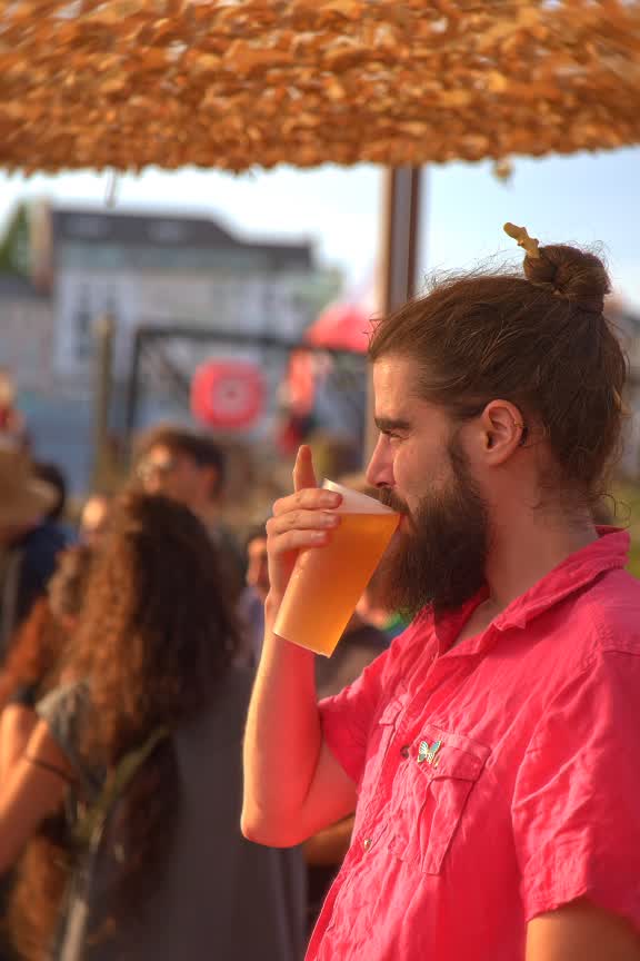 Un spectateur barbu en chemise rose boit une bière à la lumière du soir, sous un parasol de paille.