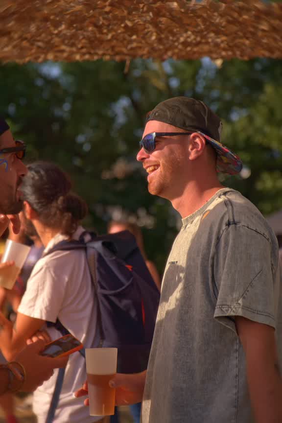 Un spectateur souriant en casquette et lunettes de soleil, un verre à la main, sous un parasol de paille.